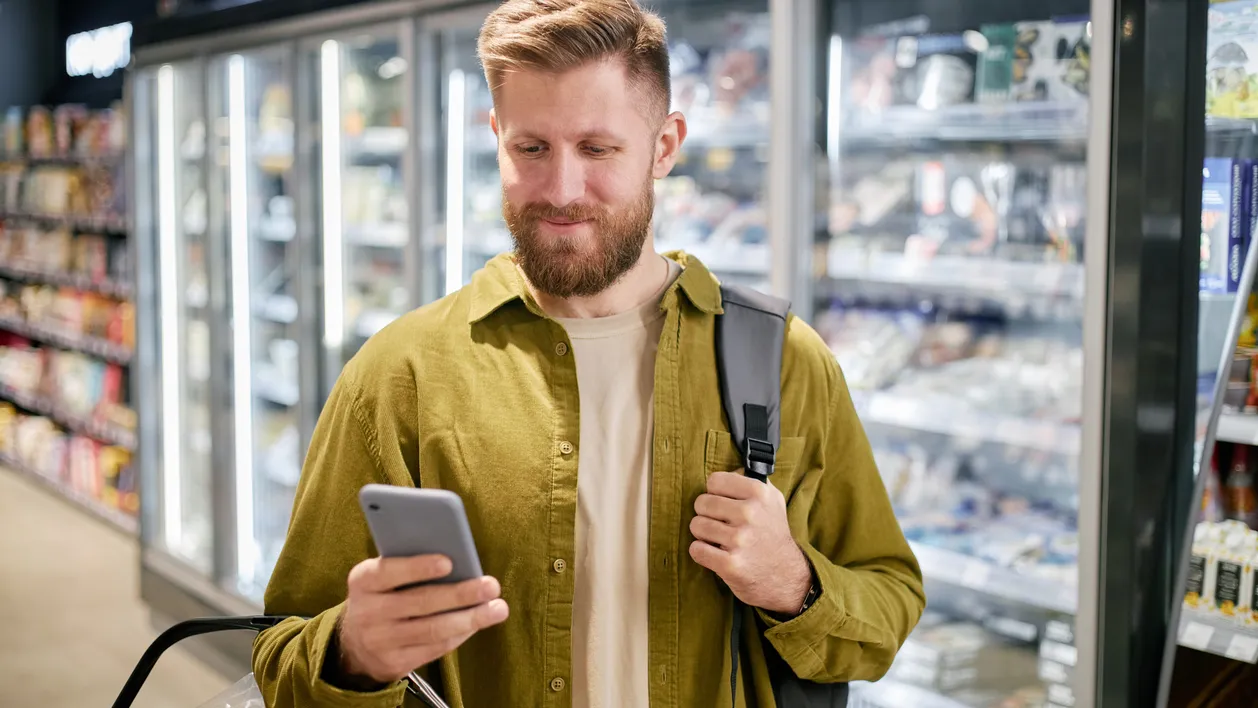 Man holding phone and shopping basket while checking grocery list in supermarket. Background shows refrigerated section with various food items and frozen goods