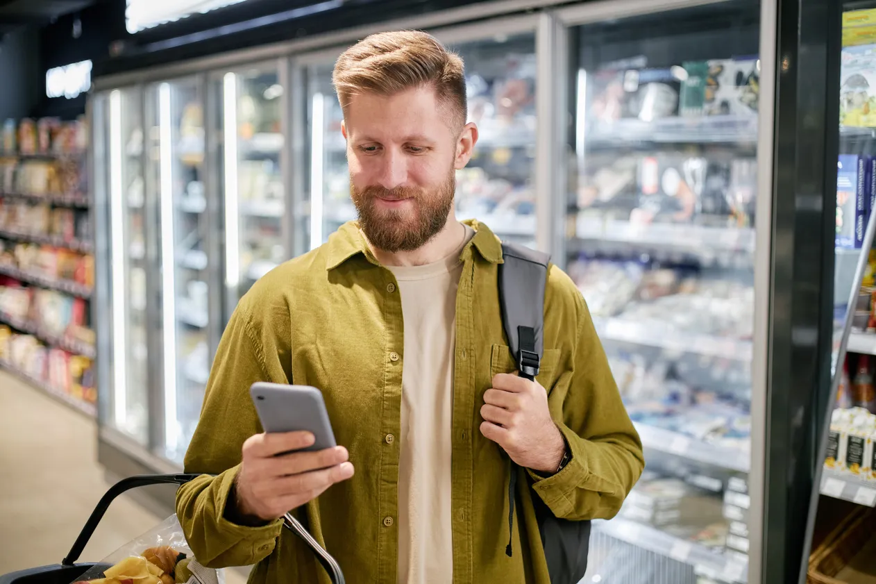 Man holding phone and shopping basket while checking grocery list in supermarket. Background shows refrigerated section with various food items and frozen goods