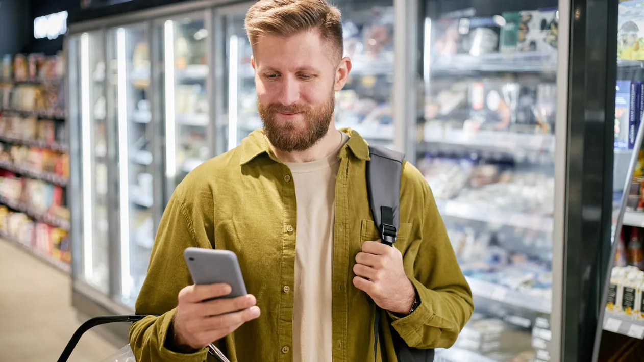 Man holding phone and shopping basket while checking grocery list in supermarket. Background shows refrigerated section with various food items and frozen goods