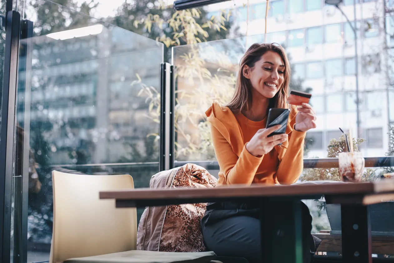 Young woman is buying online and paying with her credit card. She is sitting at the café, and enjoying her day alone. Because it is COVID-19 epidemic, she decided to sit alone. On table is protective face mask and knit hat she wore when she walked out of house.
