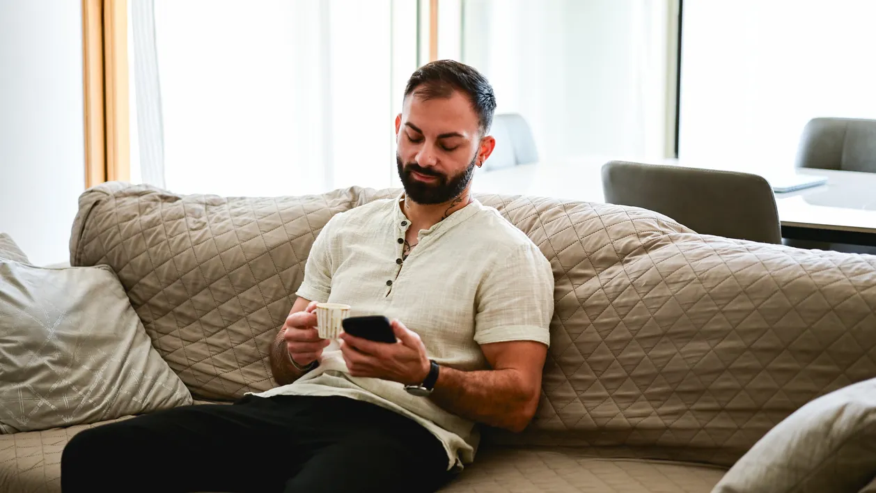 Man relaxing at home, enjoying a coffee and browsing his mobile phone