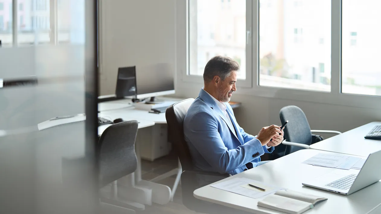 Happy busy middle aged business man investor wearing suit sitting in office using mobile phone. 50 years old smiling businessman professional banker holding smartphone at work. Authentic candid shot