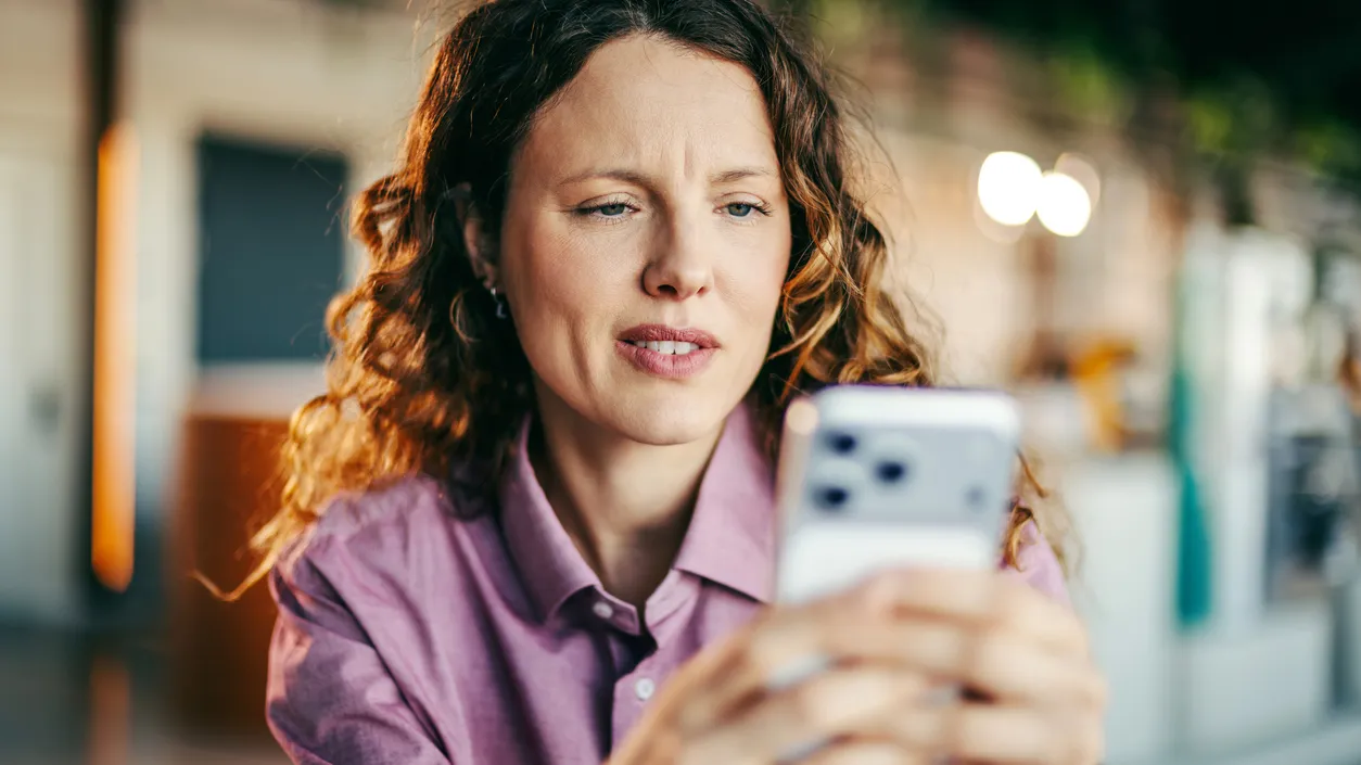 Caucasian woman with curly hair looking at her smartphone with a worried and confused expression, likely experiencing a technical problem or bad news on her mobile device