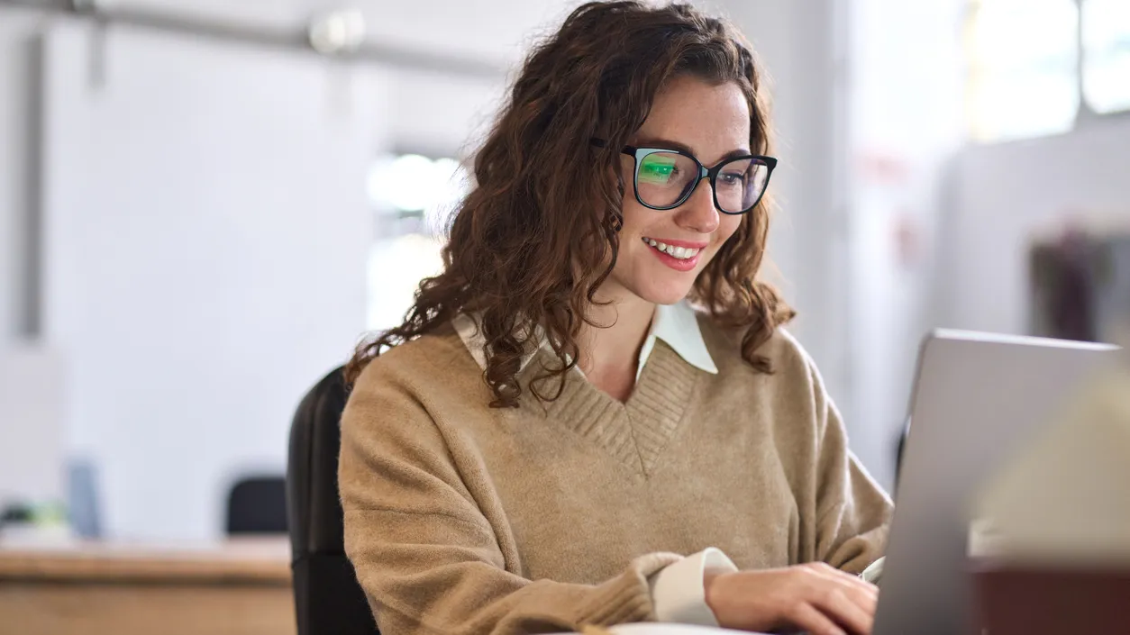 Young happy professional business woman worker employee sitting at desk working on laptop in corporate office. Smiling female student using computer technology learning online, doing web research.