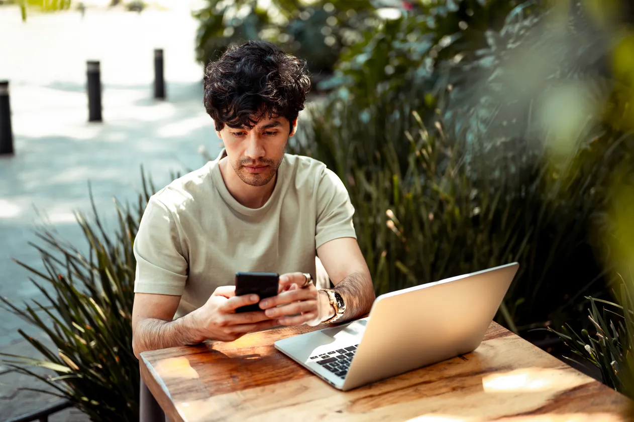 Young man working from coffee shop. He is using mobile phone and laptop