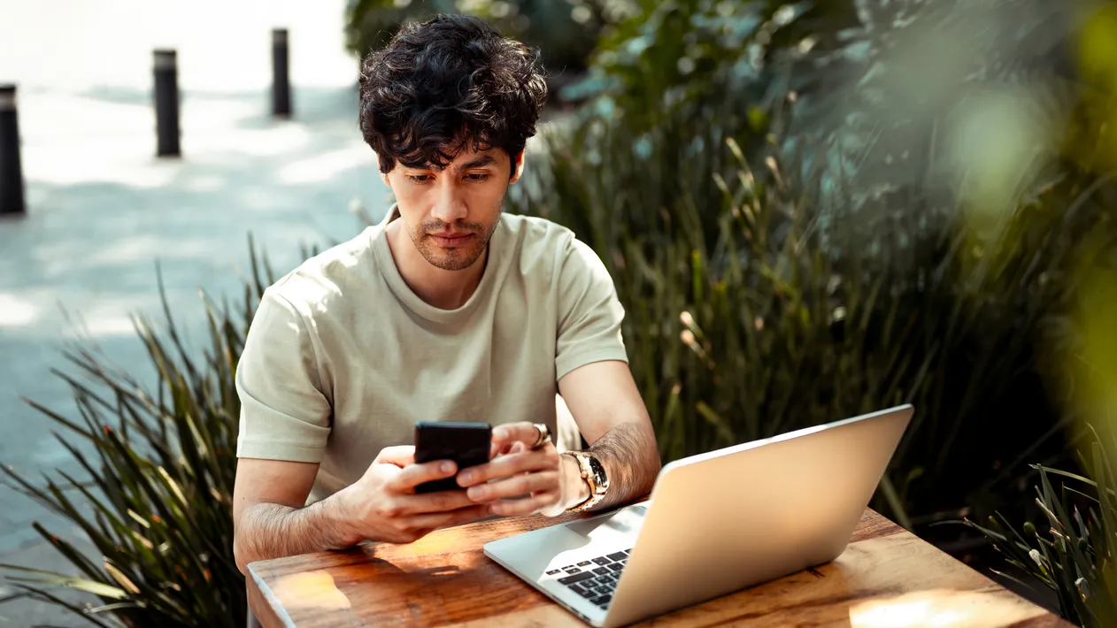 Young man working from coffee shop. He is using mobile phone and laptop