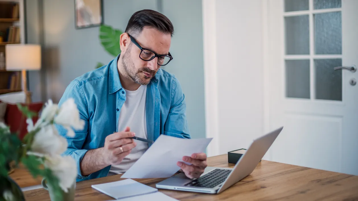 A man reading documents, sitting in front of a laptop at home.
