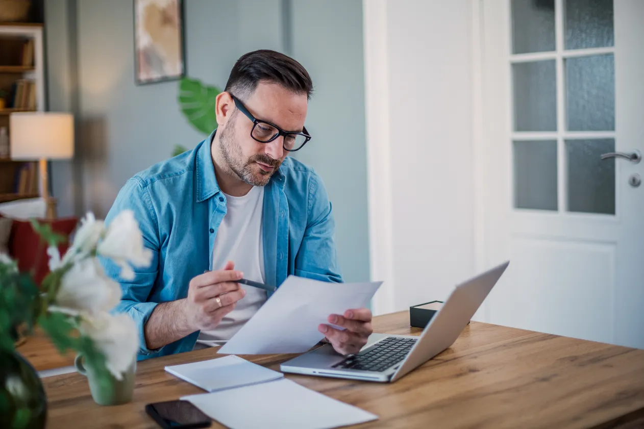 A man reading documents, sitting in front of a laptop at home.