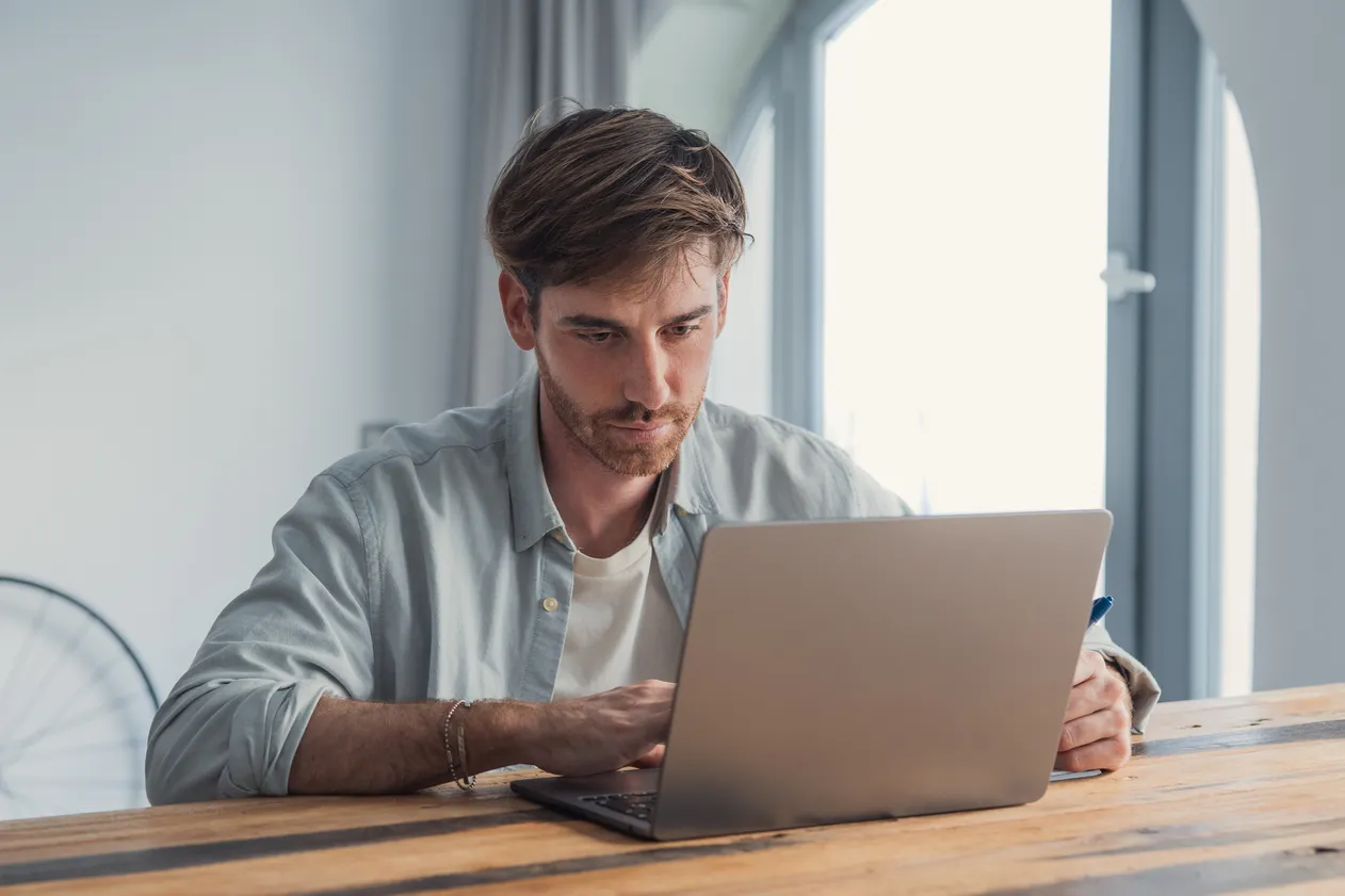 Young adult man focused at a rustic wooden desk, typing on a laptop in a bright home office, working remotely—professional, thoughtful, and productive in a modern workspace