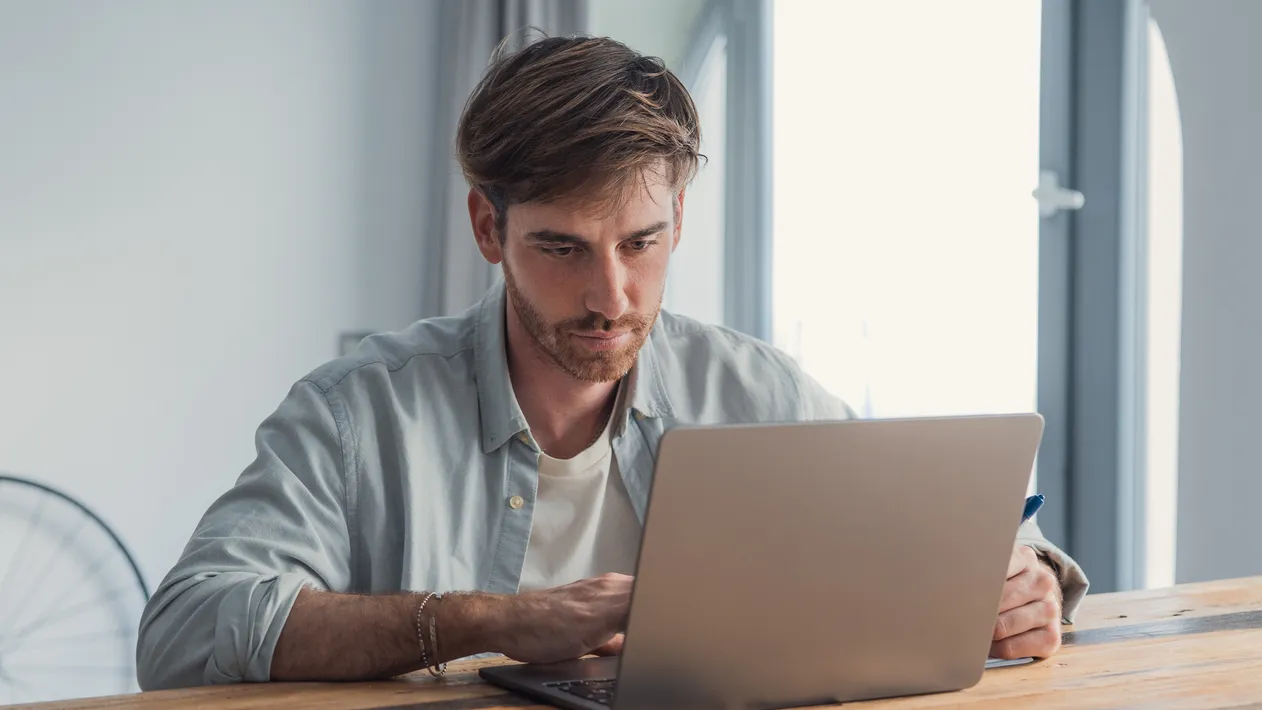 Young adult man focused at a rustic wooden desk, typing on a laptop in a bright home office, working remotely—professional, thoughtful, and productive in a modern workspace
